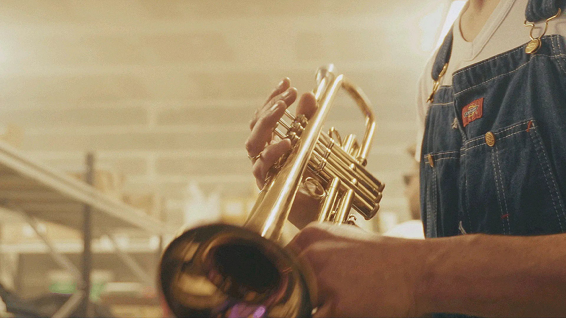 Gros plan sur un musicien jouant de la trompette en laiton, vêtu d'une salopette en jean, dans un environnement chaleureux et lumineux.