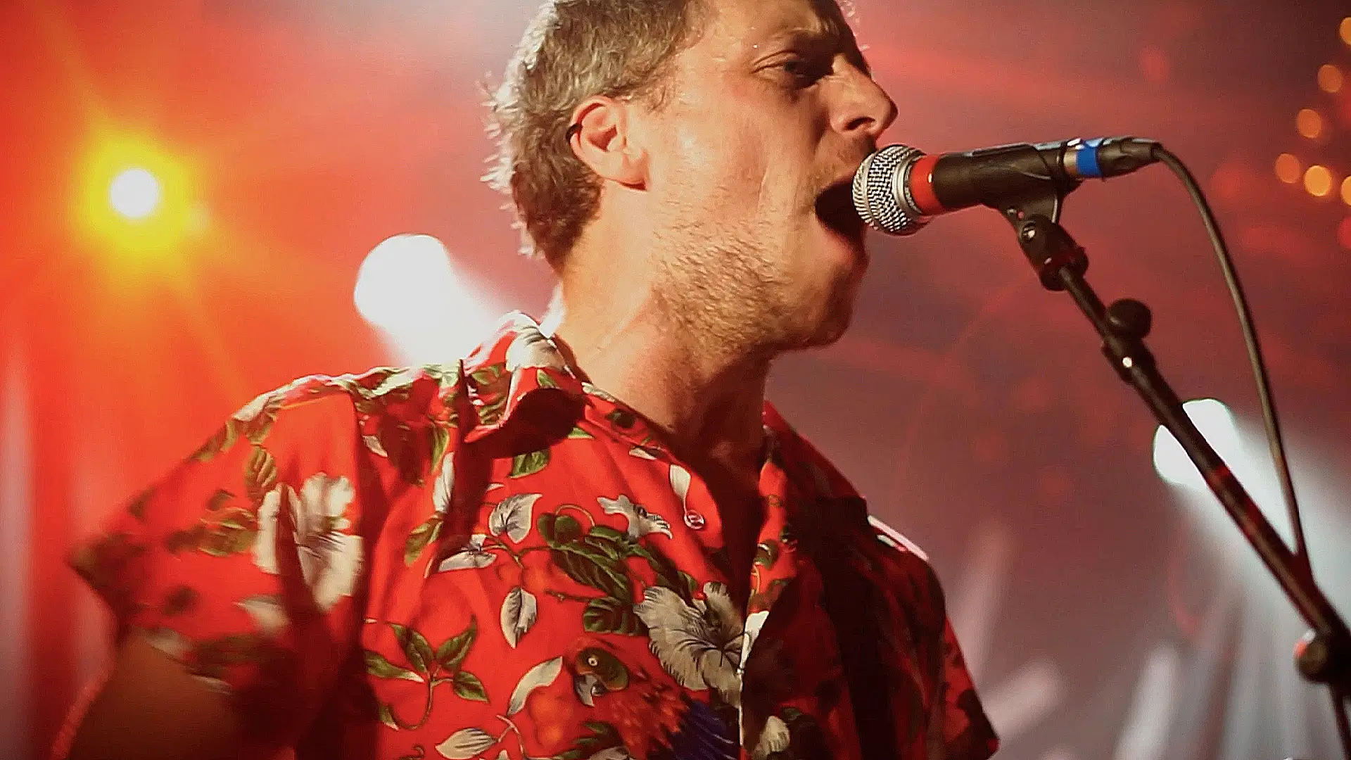 Un membre du groupe Stinky en pleine performance vocale au Ferrailleur, vêtu d'une chemise rouge à motifs tropicaux, sous des lumières de scène intenses.