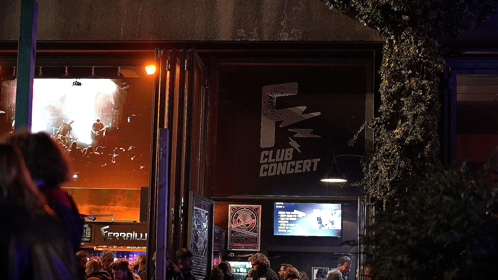 Vue extérieure du Ferrailleur, avec des spectateurs rassemblés devant l'entrée et une scène illuminée visible à travers la vitre, mettant en avant l'énergie d'une soirée de concert.