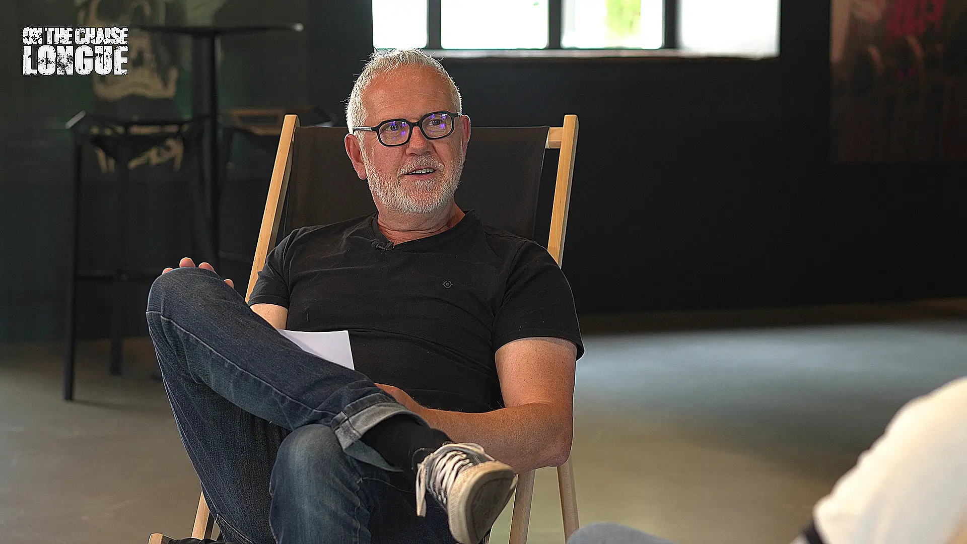 Laurent Charliot, journaliste et figure du rock français, assis sur une chaise longue avec des notes en main, interviewant dans une ambiance détendue dans les locaux du Hellfest.
