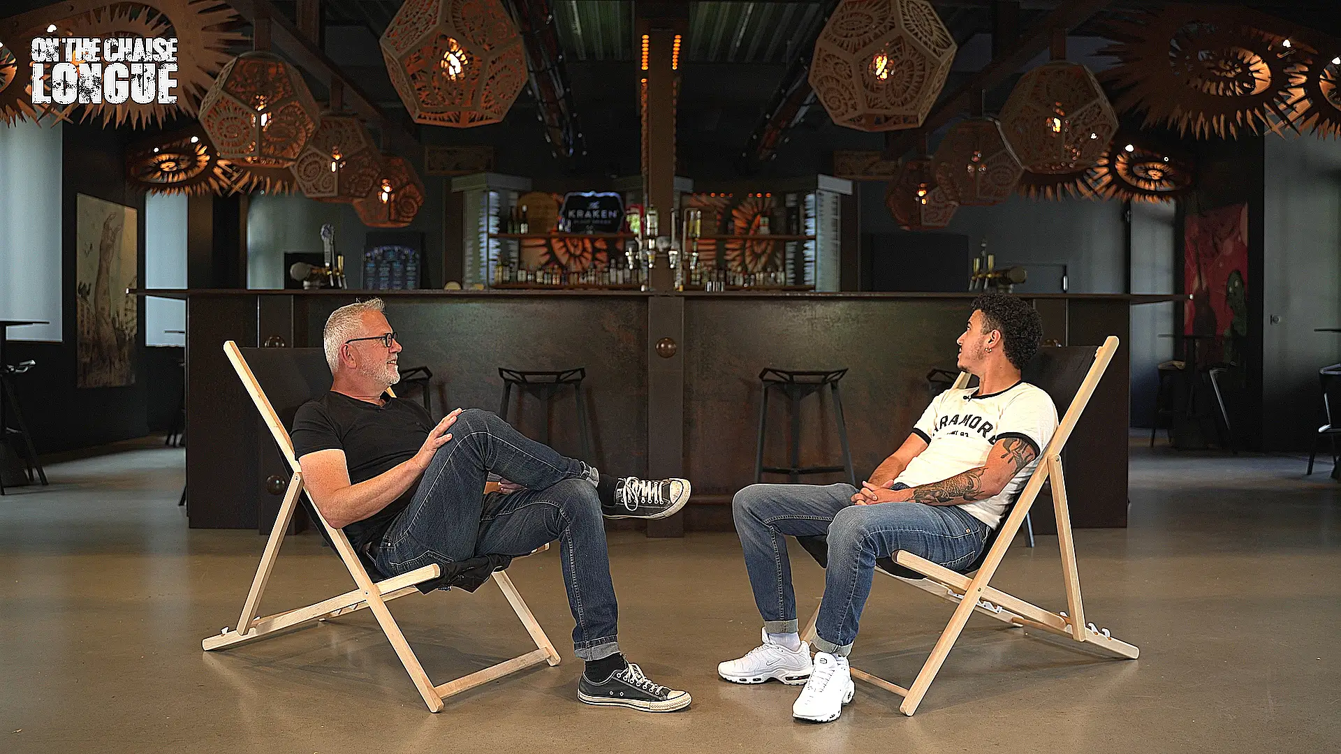 Le chanteur du groupe Stinky et Laurent Charliot, journaliste et figure du rock français, assis sur des chaises longues dans les locaux du Hellfest, discutant dans une ambiance décontractée.
