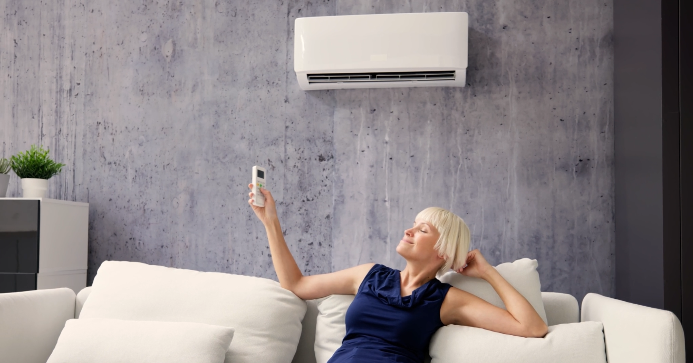 Femme allongée sur un canapé blanc, détendue, tenant une télécommande devant un climatiseur mural dans un intérieur moderne.